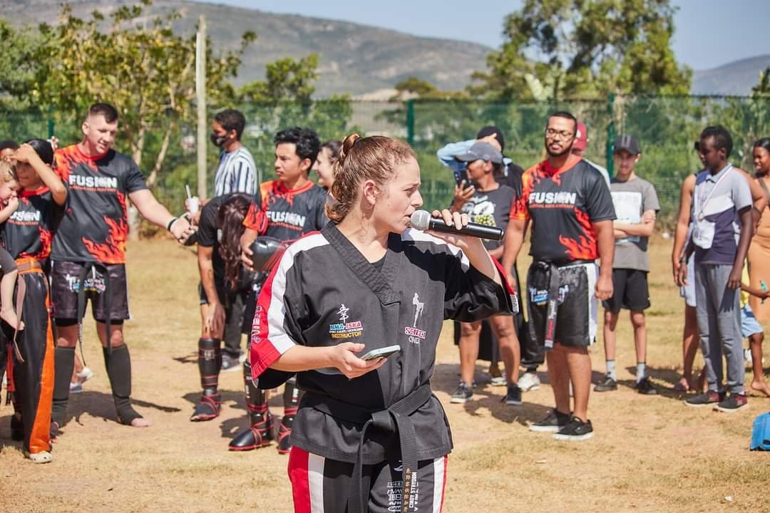 Cindy Strydom addressing participants at a Fusion Martial Arts Academy community event, microphone in hand